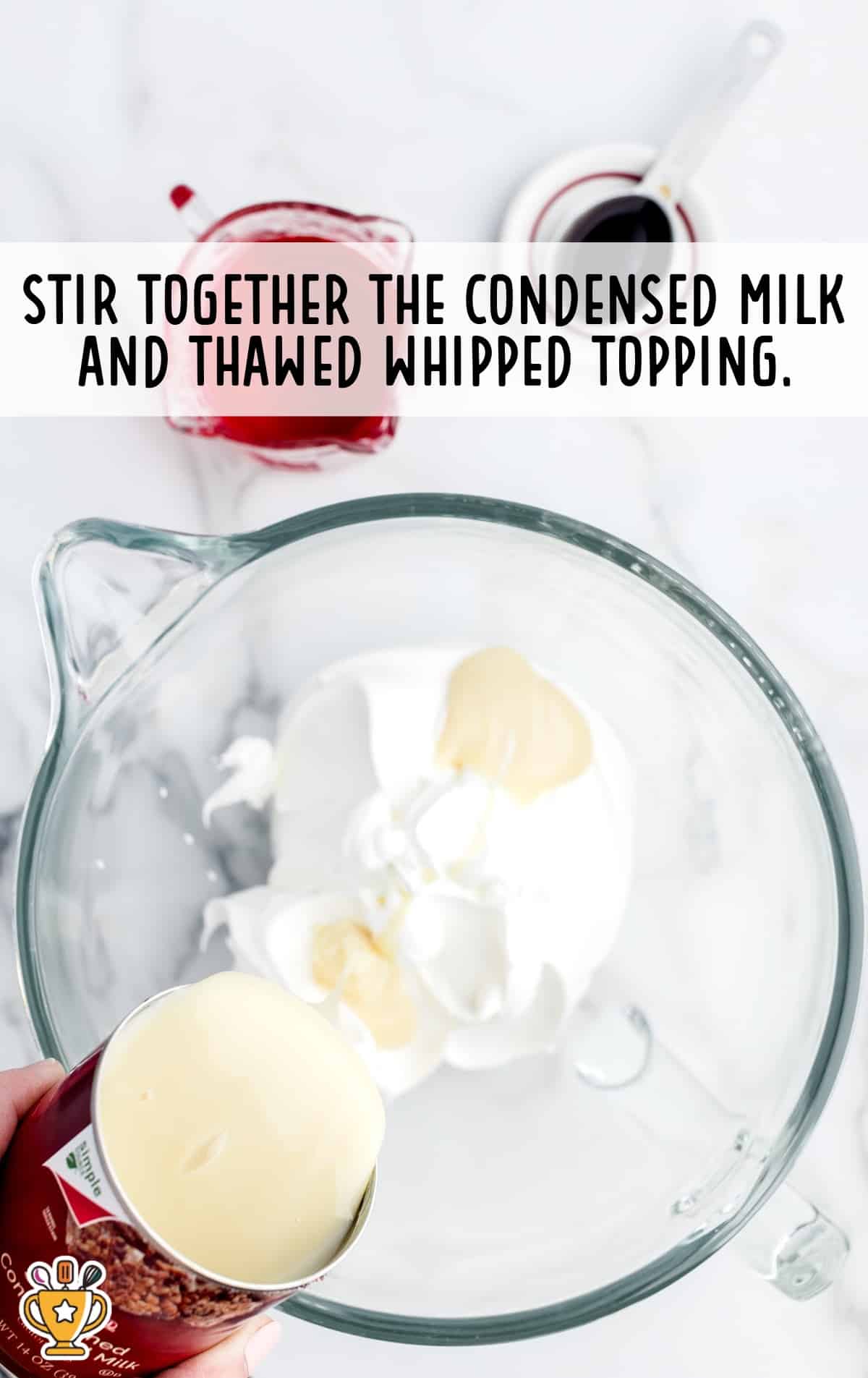 Top view of a mixing bowl with whipped topping and condensed milk being combined, surrounded by ingredients including strawberry syrup and vanilla.
