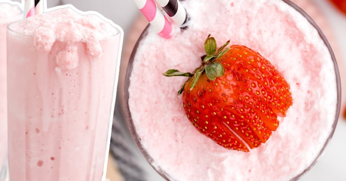 Overhead and side view of a frosty pink strawberry milkshake topped with a sliced fresh strawberry, with striped straws inside the glass.