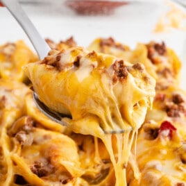 A single cheesy stuffed shell filled with seasoned taco meat being lifted from a tray, with stretchy melted cheese trailing beneath.