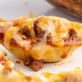 Single taco stuffed shell on a white plate, loaded with beef and melted cheese, with more shells and taco sauce blurred in the background.