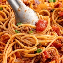 Close-up of spaghetti mixed with ground beef, diced tomatoes, bacon, and green onions, being twirled with metal tongs.