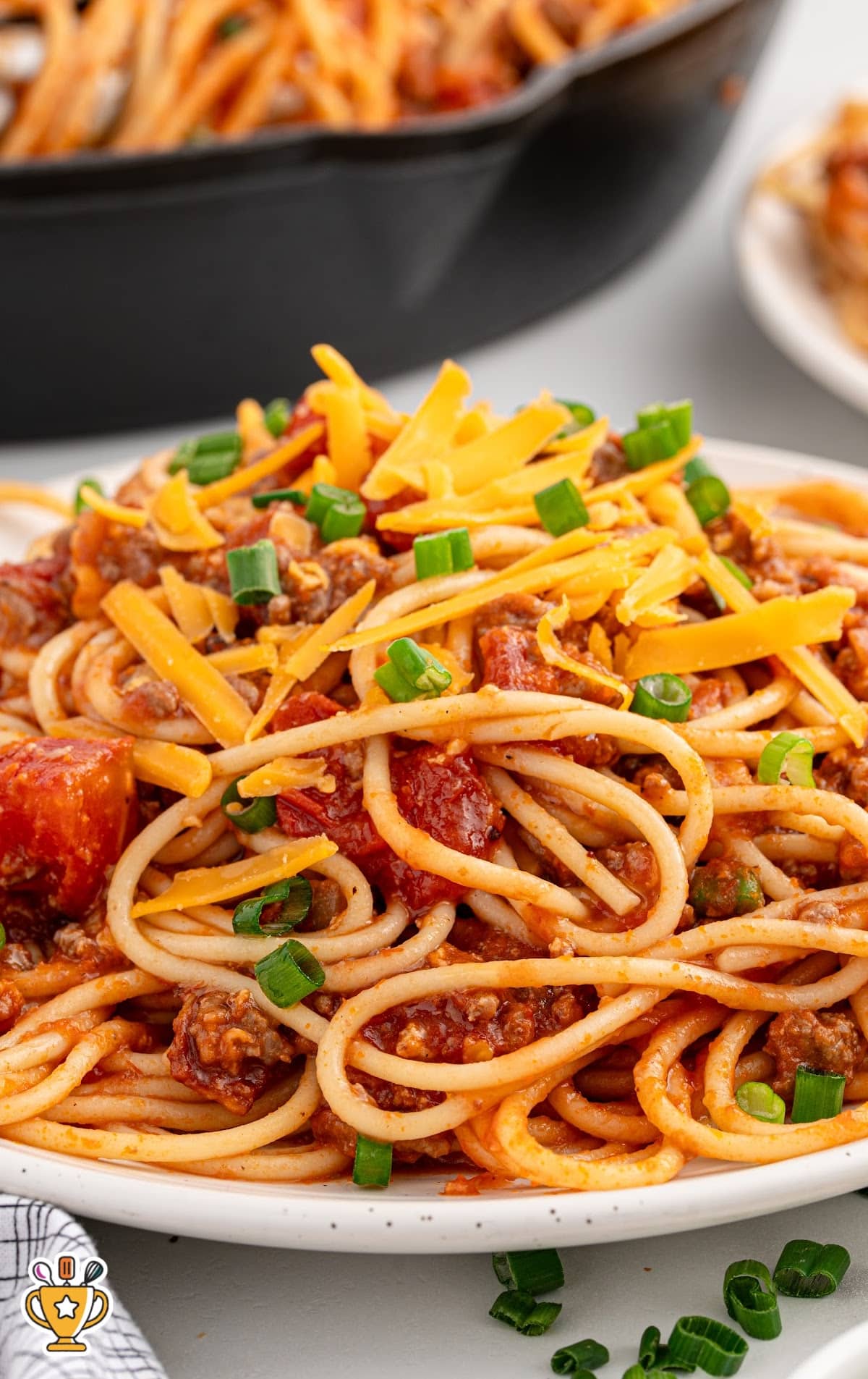 A plate of cowboy spaghetti topped with shredded cheddar and green onions, with a skillet of pasta blurred in the background.