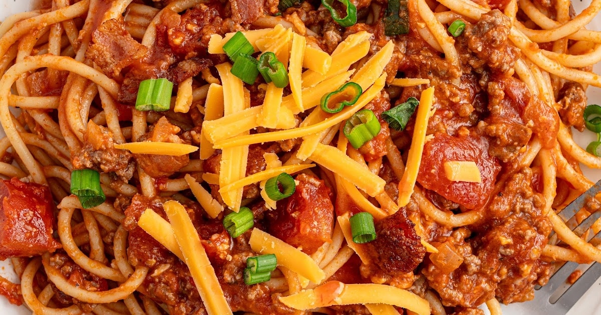 Overhead close-up of cowboy spaghetti topped with melted cheddar cheese, chopped green onions, ground beef, and thick tomato chunks.
