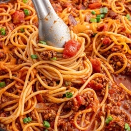 Cowboy spaghetti in a skillet being swirled with tongs, with visible bacon, tomatoes, and green onions in a cheesy tomato sauce.