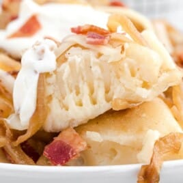 Close-up of crispy fried pierogies topped with caramelized onions, bacon bits, shredded cheese, and a dollop of sour cream on a white plate.