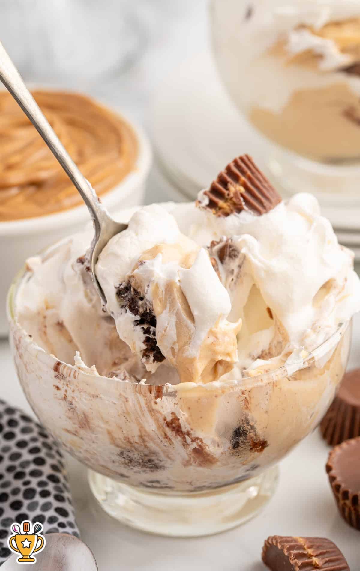 Close-up of a serving bowl with layers of whipped topping, peanut butter mousse, brownies, and peanut butter cups, ready to be eaten with a spoon.