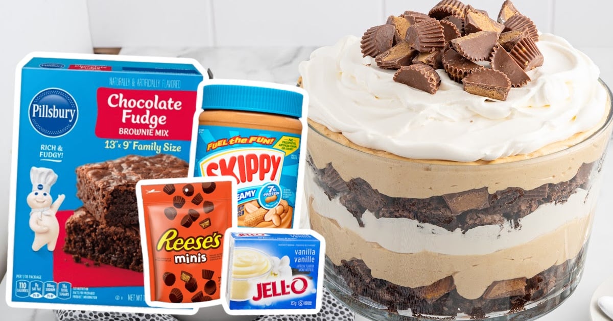 Large glass trifle bowl topped with peanut butter cups surrounded by ingredient packaging including brownie mix, Skippy peanut butter, Reese’s minis, and Jell-O pudding.