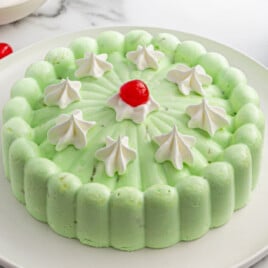 Whole seafoam salad molded into a scalloped ring on a white plate, pale green with whipped cream dollops and a maraschino cherry in the center. Surrounded by extra cherries, stacked white plates with forks, and a checkered napkin on the side.