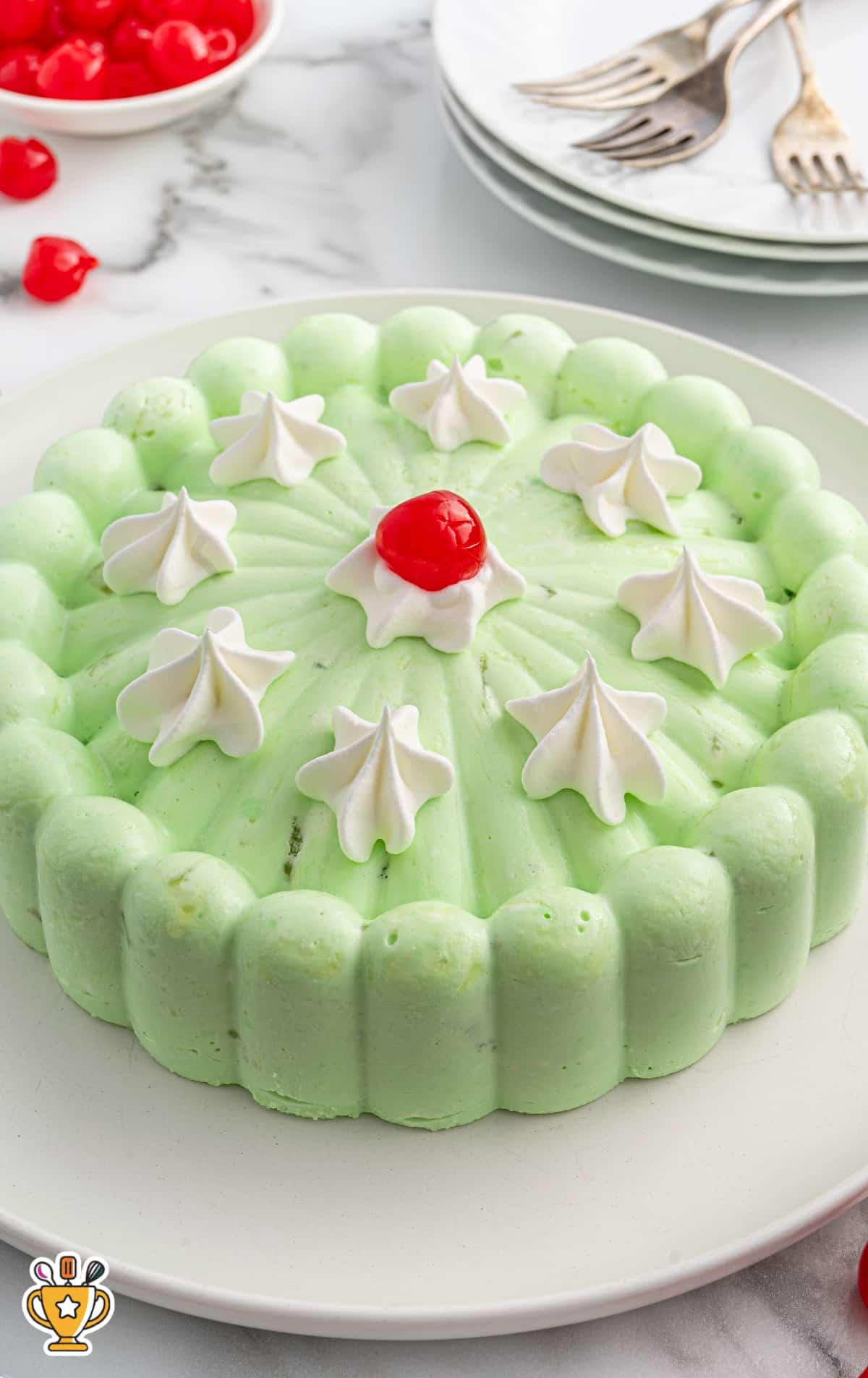 Overhead view of the full molded seafoam salad garnished with dollops of whipped cream and a cherry in the center, with stacked plates and forks nearby.