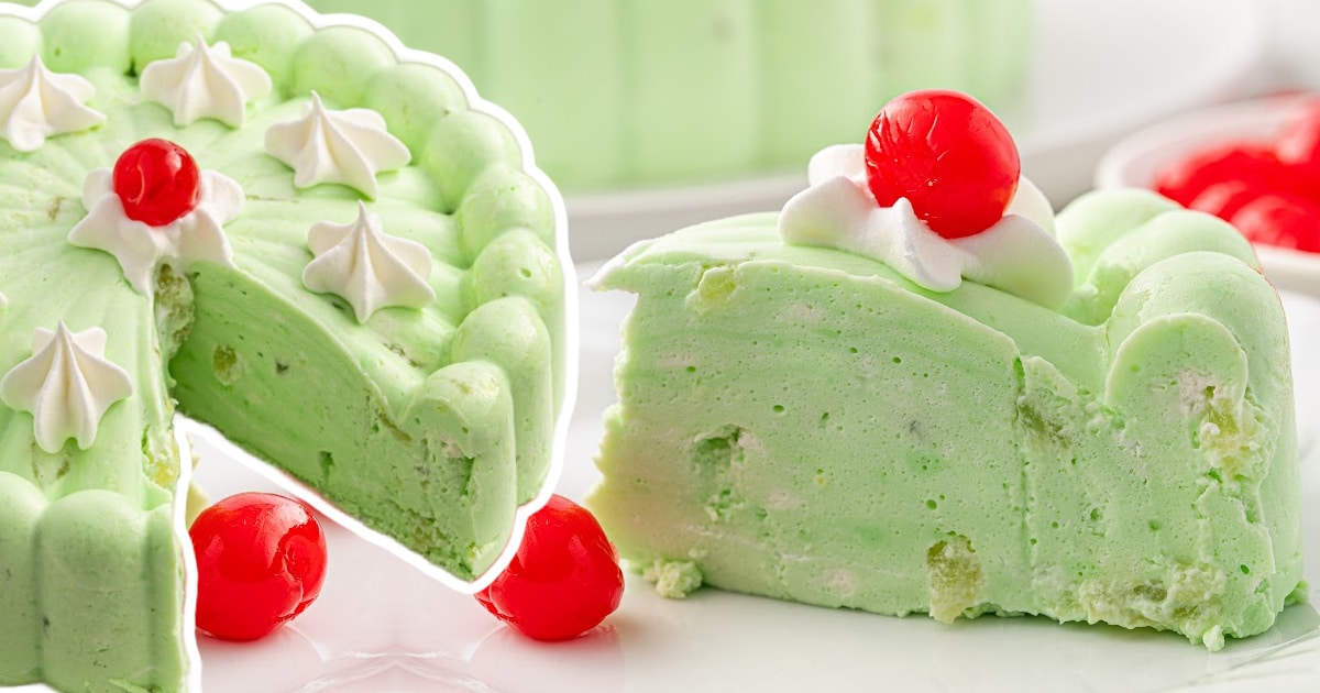 Wide view of a seafoam salad slice on a white plate, with whipped cream and a cherry on top and a bowl of cherries behind.