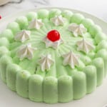Molded seafoam salad on a white plate, light green in color with a scalloped edge. Topped with dollops of whipped cream and a single red maraschino cherry in the center for garnish.