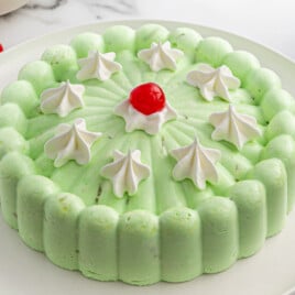 Molded seafoam salad on a white plate, light green in color with a scalloped edge. Topped with dollops of whipped cream and a single red maraschino cherry in the center for garnish.