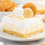 A square slice of banana cheesecake bar on a white plate, with a golden vanilla wafer crust, creamy cheesecake filling, and a thick whipped topping. The bar is garnished with a swirl of whipped cream, a banana slice, and a vanilla wafer cookie, with more wafers blurred in the background.