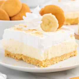 A square slice of banana cheesecake bar on a white plate, with a golden vanilla wafer crust, creamy cheesecake filling, and a thick whipped topping. The bar is garnished with a swirl of whipped cream, a banana slice, and a vanilla wafer cookie, with more wafers blurred in the background.