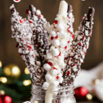 A festive mason jar filled with white and dark chocolate-covered pretzel sticks, coated in crushed peppermint and mini marshmallows, arranged upright for display. A decorative tin filled with additional chocolate stir sticks sits beside it, with Christmas ornaments and lights in the blurred background.
