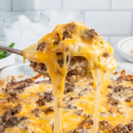 A close-up of a spoonful of cheesy hamburger potato casserole being lifted from the baking dish, creating a long, gooey cheese pull.