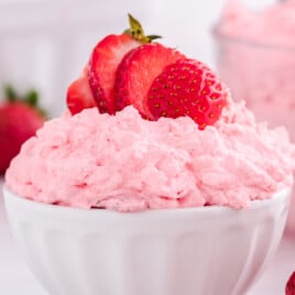 A white scalloped bowl filled with fluffy pink strawberry jello salad, topped with fresh sliced strawberries. The creamy, whipped texture looks light and airy, with more pink salad and whole strawberries blurred in the background.