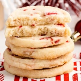 Stack of peppermint shortbread cookies with red candy bits, topped with a half-eaten cookie, surrounded by Andes peppermint candies and red holiday ornaments.
