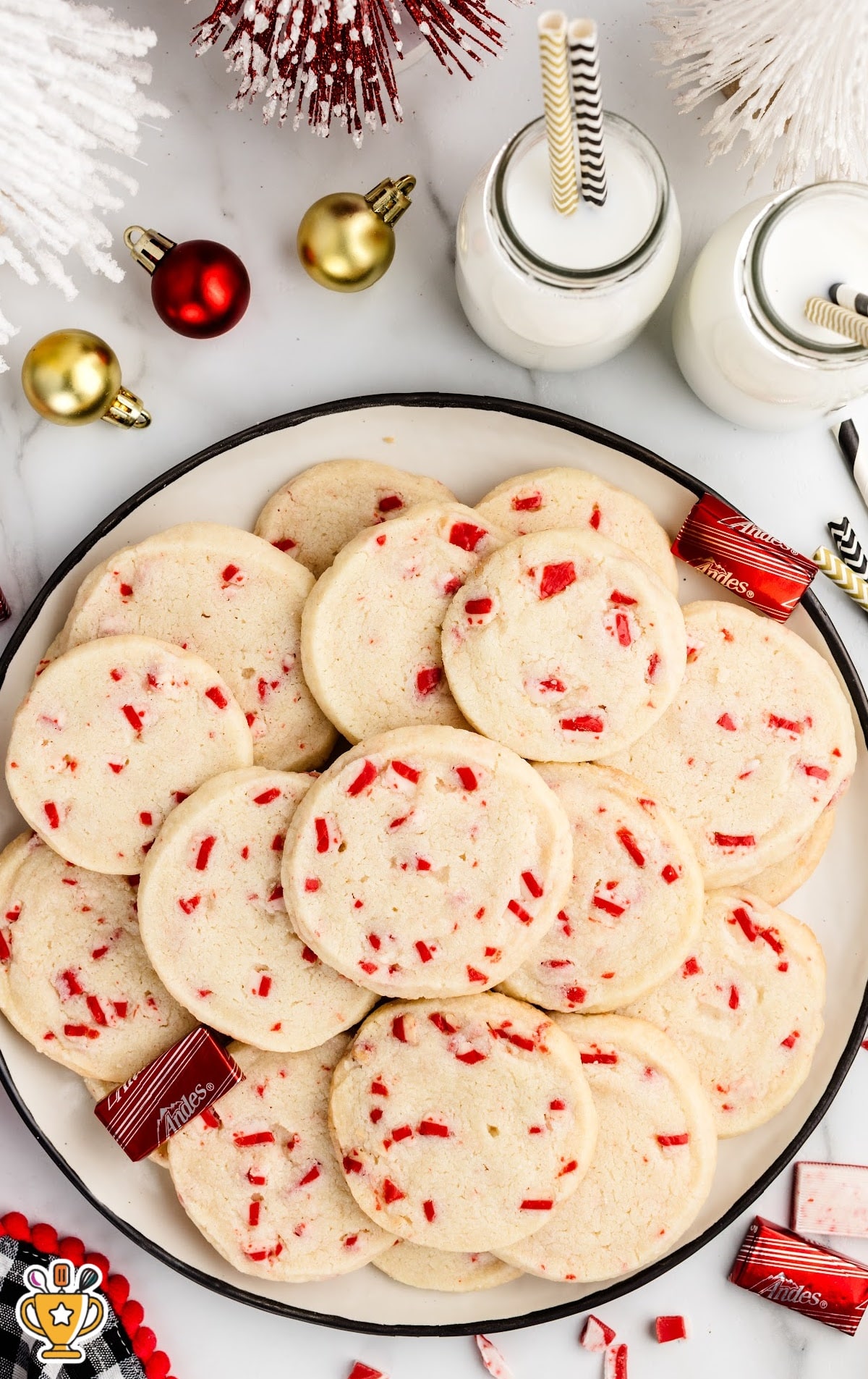 Overhead view of a plate filled with peppermint shortbread cookies, surrounded by festive red and gold ornaments, milk bottles with striped straws, and Andes peppermint candies.