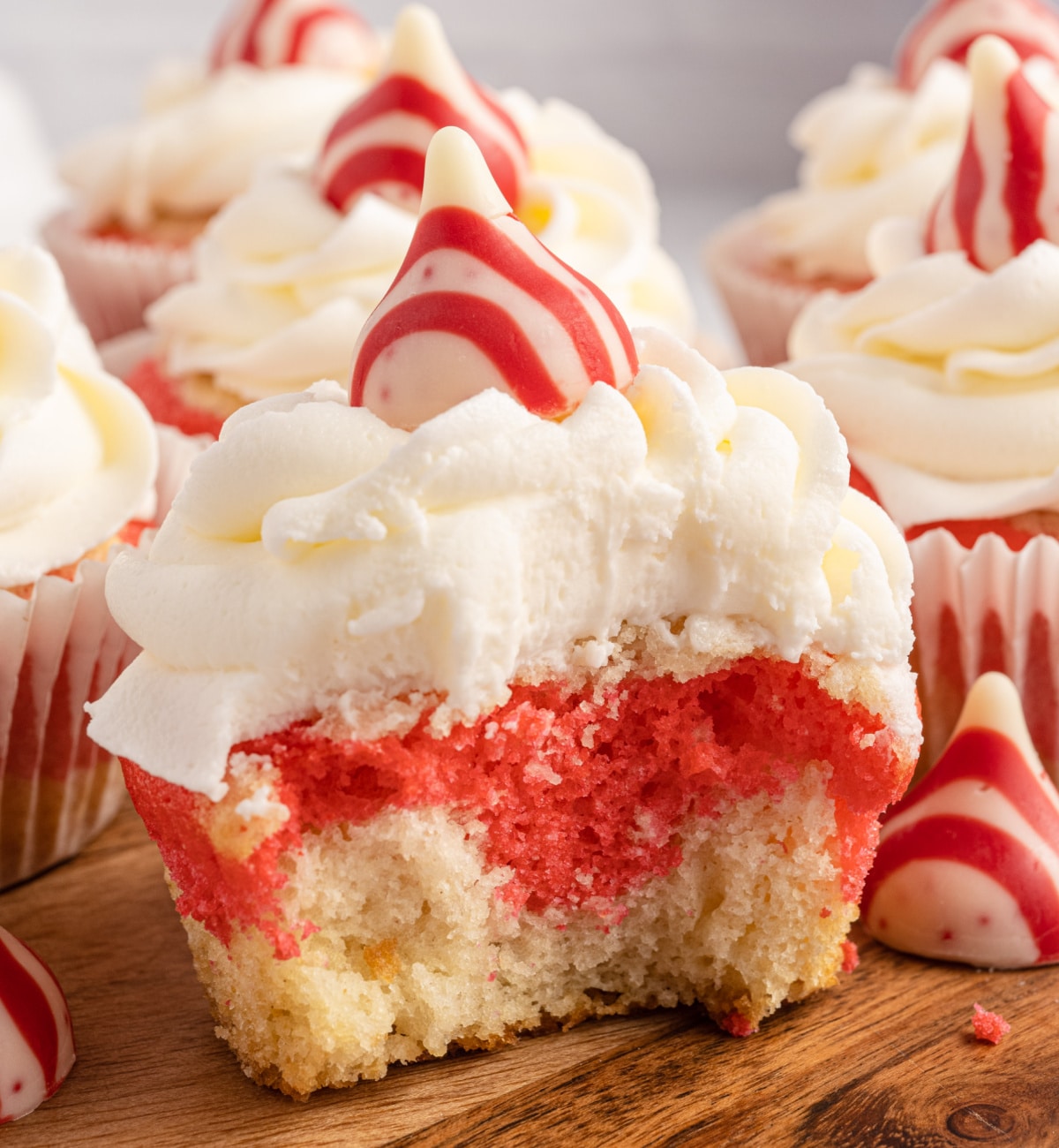 Close-up of candy cane cupcakes with swirled red and white cake layers, topped with creamy white frosting and a striped peppermint candy kiss, served on a wooden board. A delicious Candy Cane Dessert.