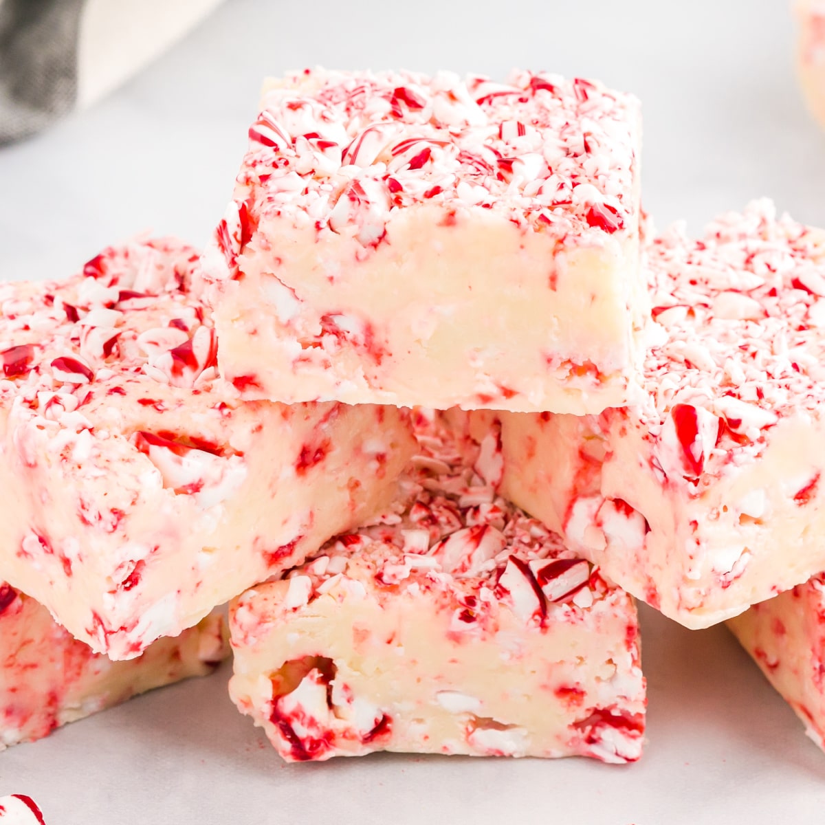 Close-up of creamy white chocolate candy cane fudge squares loaded with crushed peppermint pieces, stacked on a white surface.