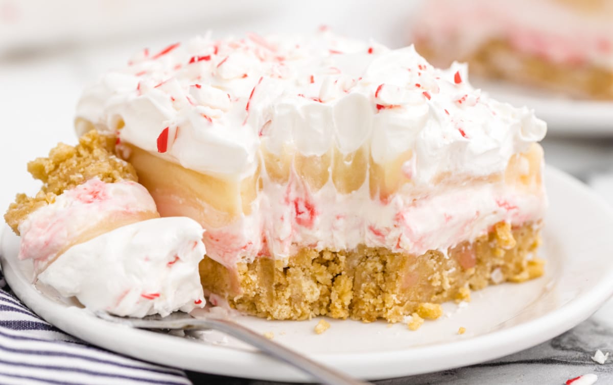 Close-up of a layered candy cane dessert with a golden graham cracker crust, creamy pudding filling, fluffy whipped topping, and crushed candy canes sprinkled on top, served on a white plate with a spoonful on the side.