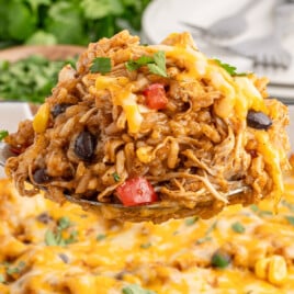 A heaping spoonful of cheesy Mexican chicken and rice casserole with shredded chicken, fluffy rice, melted cheese, black beans, corn, and red peppers, garnished with fresh cilantro, lifted above a baking dish filled with the same golden, gooey casserole.