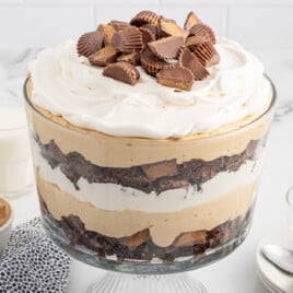 Large glass trifle bowl filled with layers of brownies, peanut butter pudding, whipped cream, and chopped peanut butter cups, topped with more Reese’s pieces. The dessert is displayed on a marble surface with bowls, spoons, a dish of peanut butter, and glasses of milk with striped straws in the background.