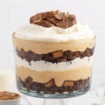Glass trifle bowl filled with layers of peanut butter mousse, whipped cream, chocolate brownies, and chopped peanut butter cups, topped with more peanut butter cup pieces on top.