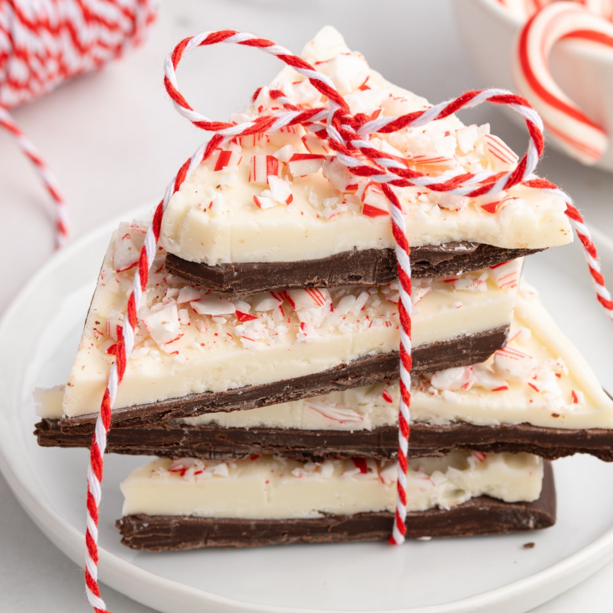 Stack of homemade peppermint bark with layers of dark and white chocolate topped with crushed candy canes, tied with red and white string on a white plate.