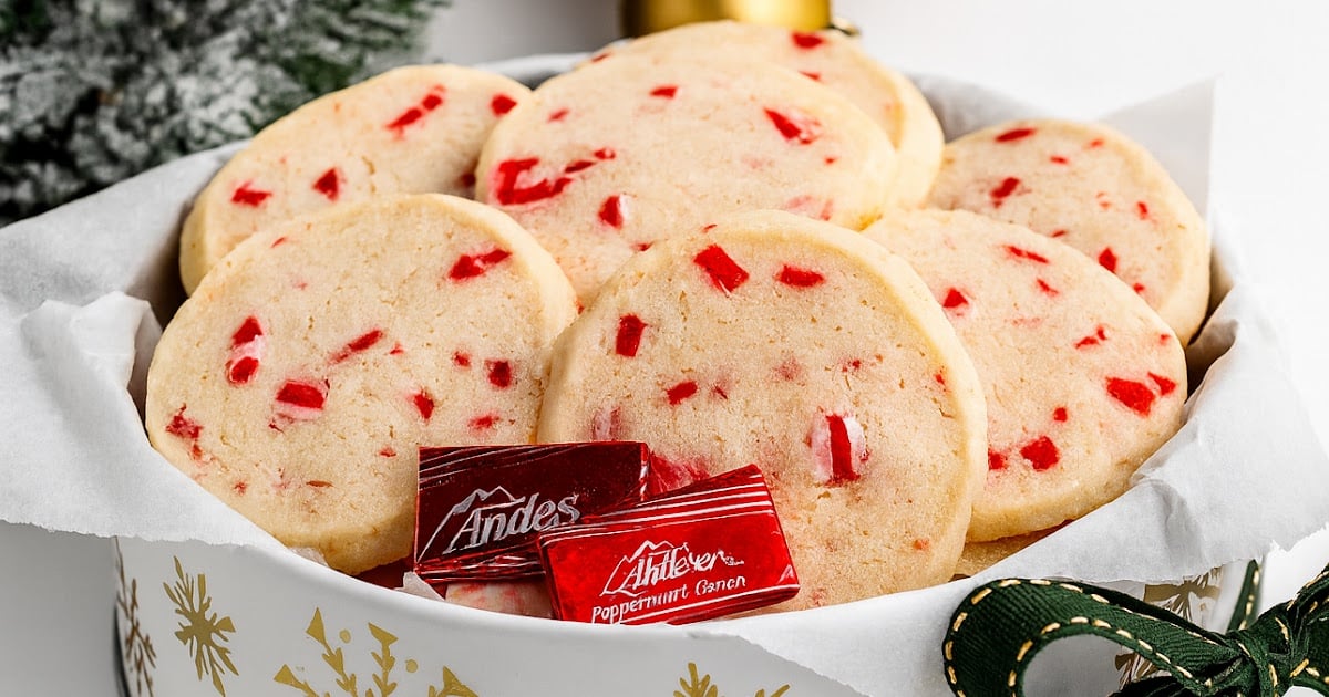 Festive tin lined with parchment and filled with peppermint shortbread cookies, garnished with Andes peppermint candies, placed near Christmas decorations and greenery.