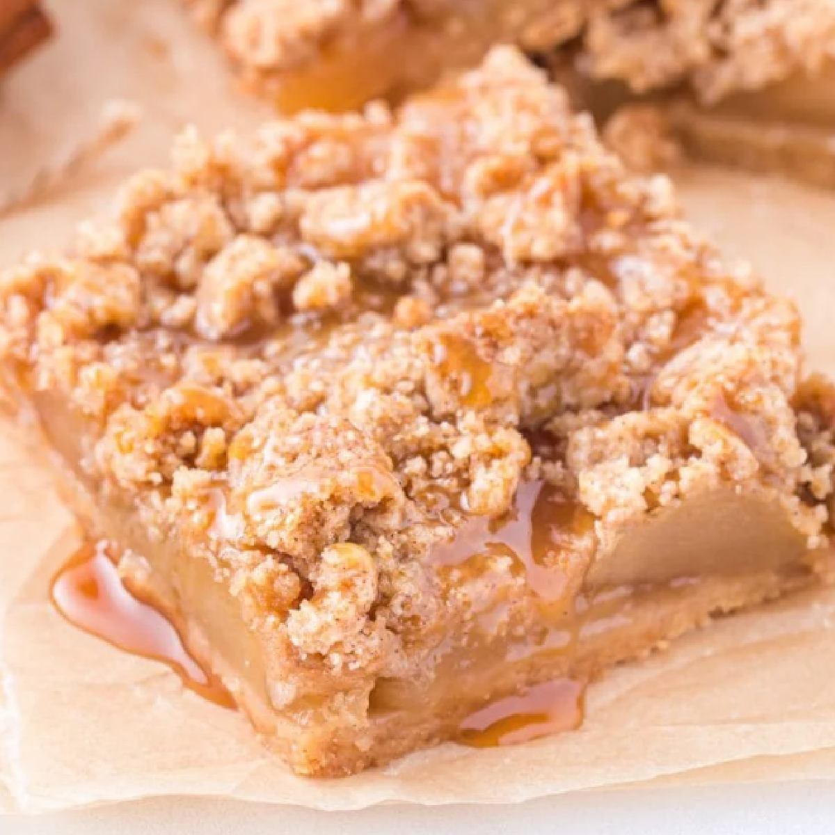 Soft and chewy caramel apple bar with a buttery crumble topping, drizzled with gooey caramel sauce and filled with tender cinnamon-spiced apple filling — served on parchment paper for a cozy fall dessert.