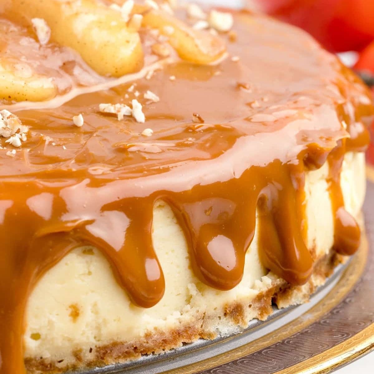 Close-up of a rich caramel apple cheesecake with a creamy baked filling, golden crust, and thick caramel sauce dripping over the edges, topped with tender cinnamon-spiced apple slices and chopped nuts.