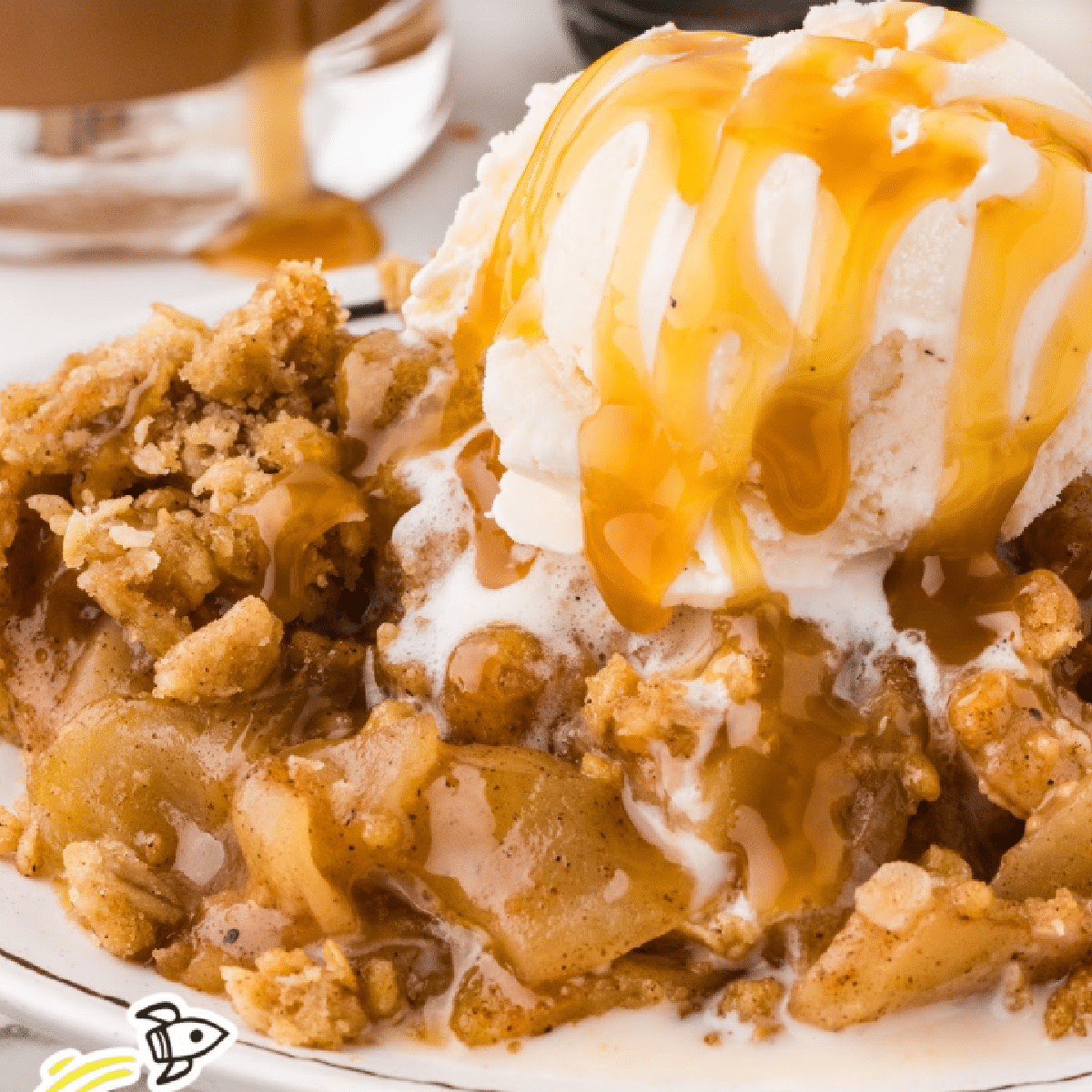 Warm caramel apple crisp topped with vanilla ice cream and drizzled with rich caramel sauce on a white plate.