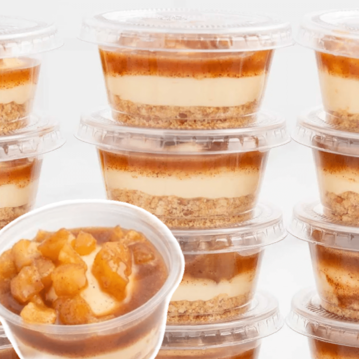 Stacks of individual caramel apple dessert cups in clear plastic containers, layered with buttery graham cracker crust, creamy cheesecake filling, and cinnamon apple pie topping — perfect no-bake fall treat for parties or gifting.