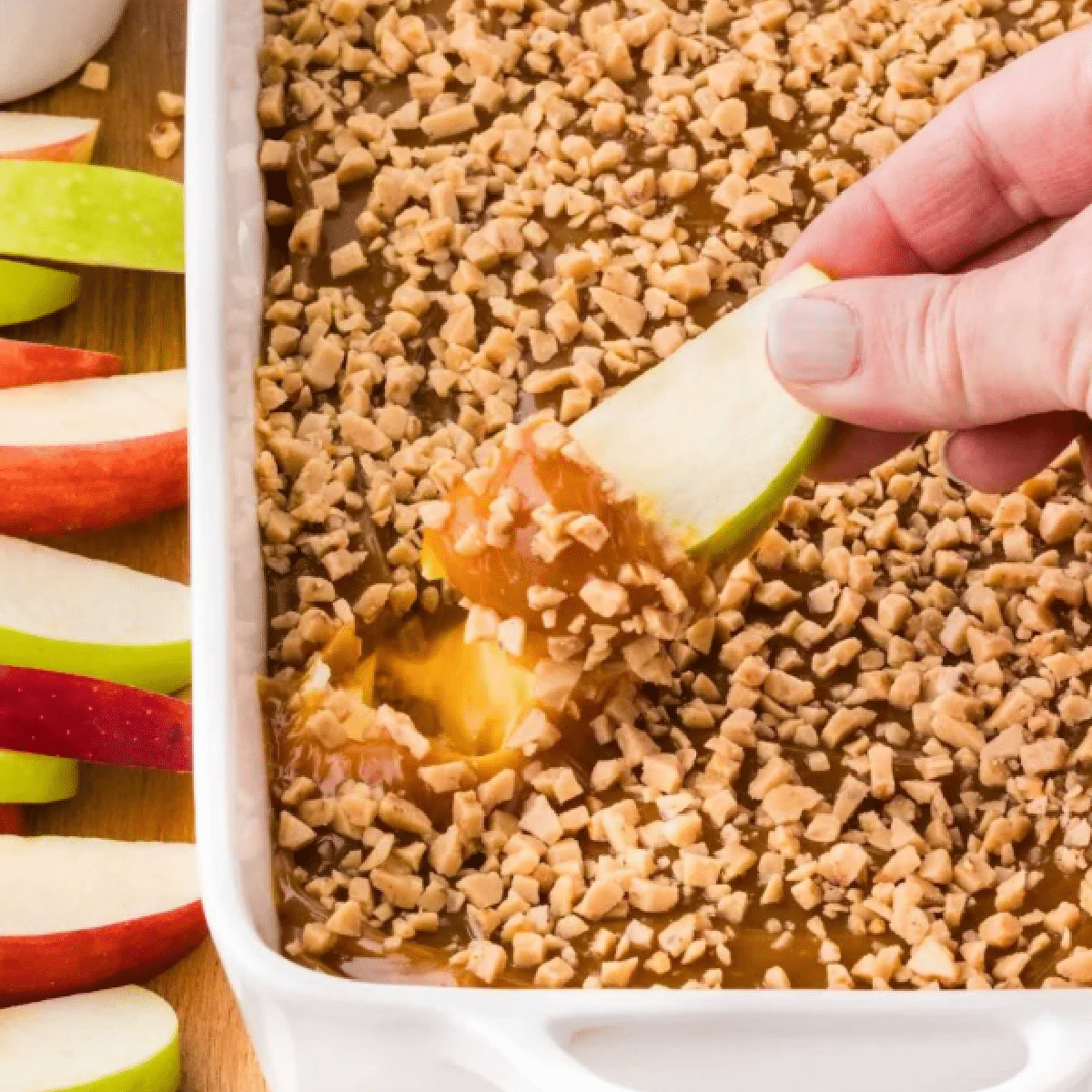 Crisp green apple slice dipped into a layered caramel apple dip topped with crunchy toffee bits, served in a white dish surrounded by red and green apple slices — easy no-bake dessert perfect for parties and fall gatherings.
