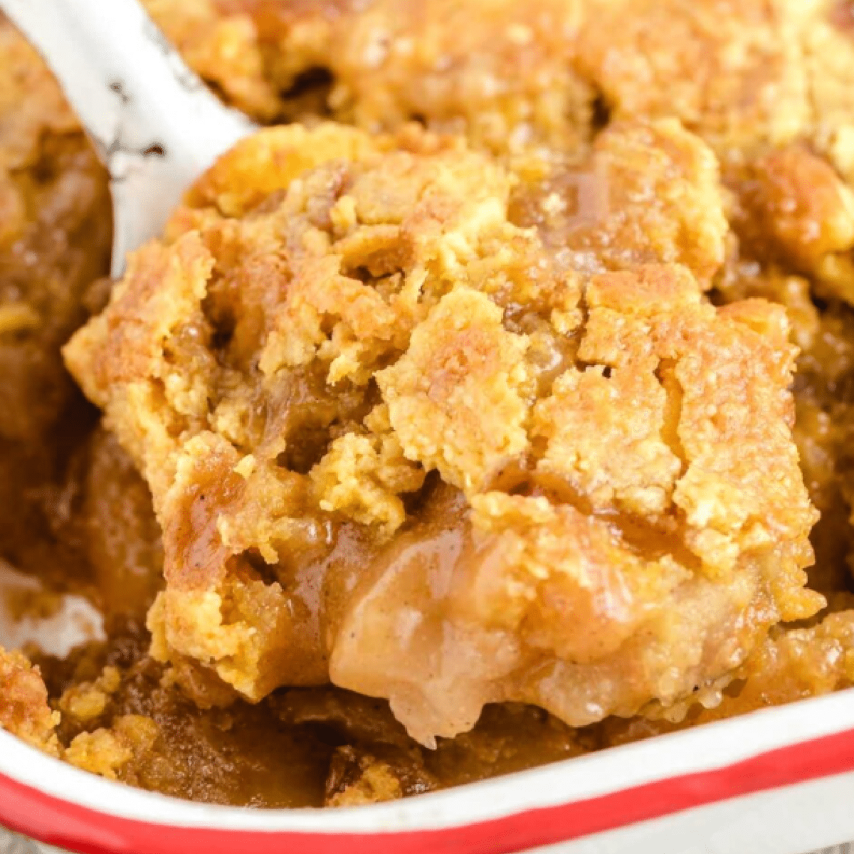 Warm serving of caramel apple dump cake scooped from a white baking dish with a red rim, showing a gooey apple filling and golden brown cake topping with caramel sauce baked in — easy fall dessert made with cake mix and pie filling.