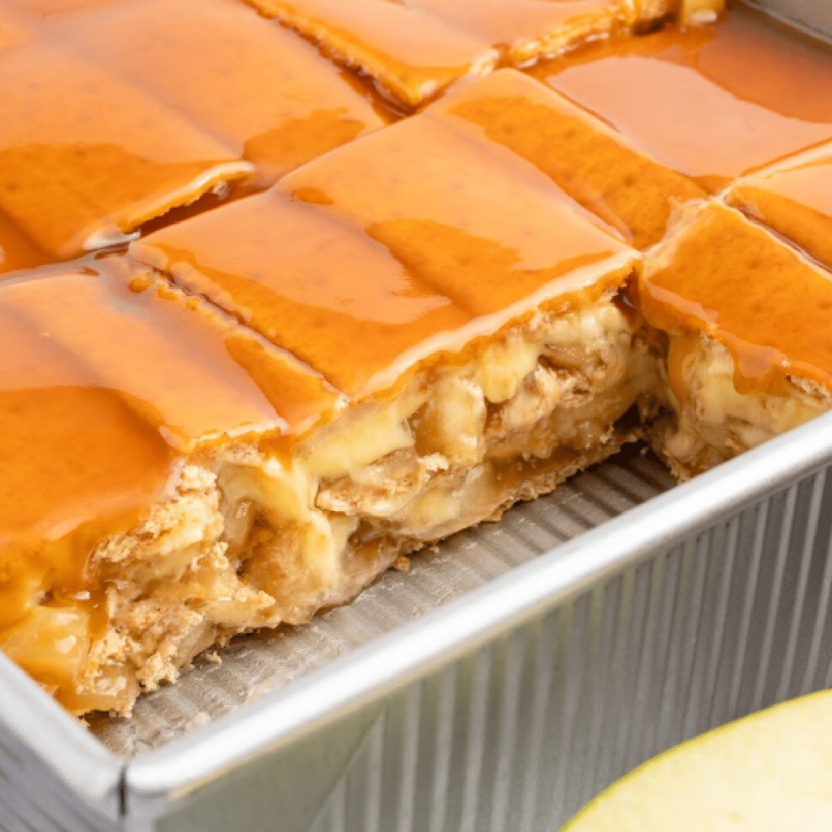 Layered caramel apple éclair cake in a metal baking pan, made with graham crackers, apple pie filling, creamy vanilla pudding, and topped with a glossy caramel glaze — no-bake dessert sliced and ready to serve.