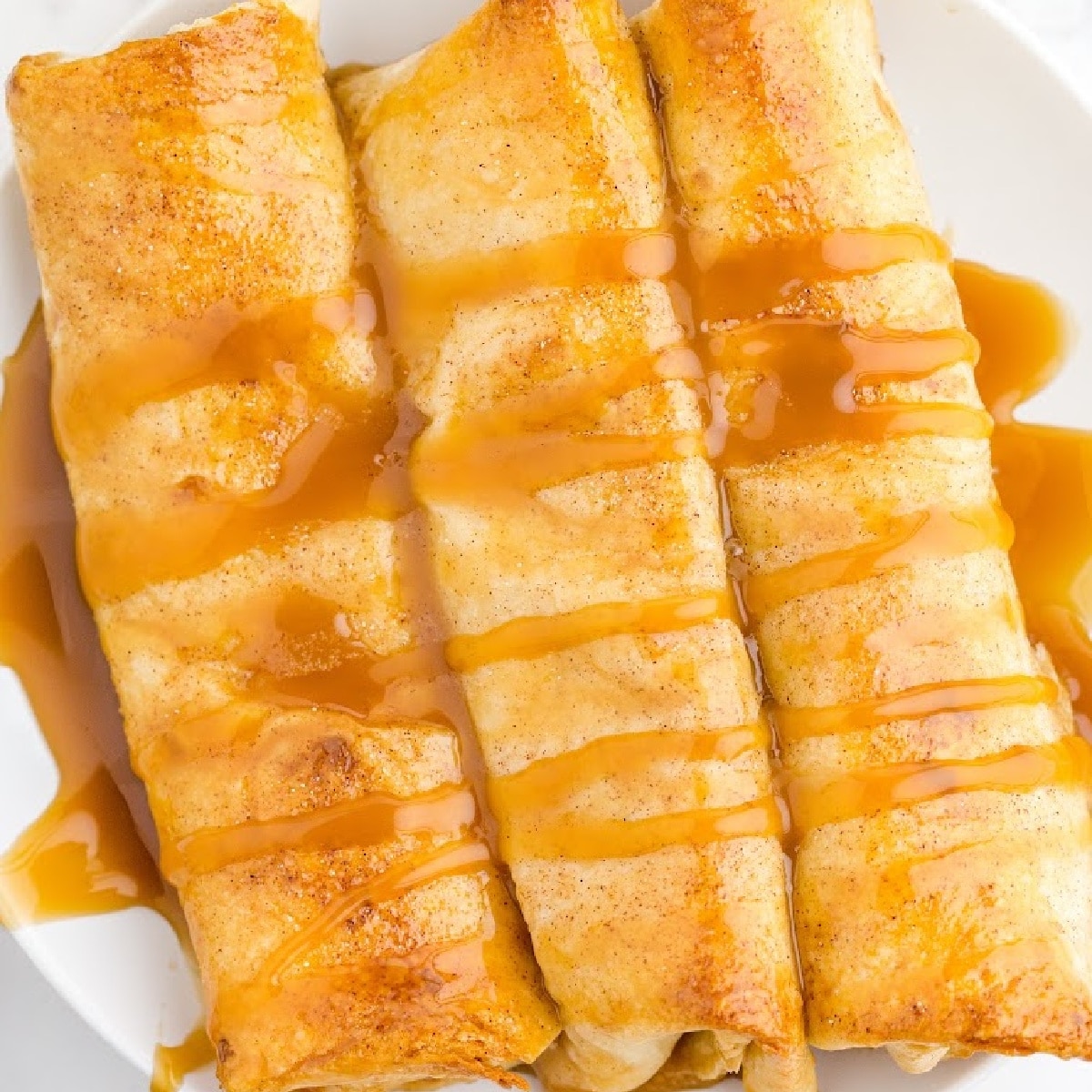 Three golden-brown caramel apple enchiladas on a white plate, drizzled generously with warm caramel sauce, featuring soft cinnamon tortillas wrapped around a sweet apple pie filling.