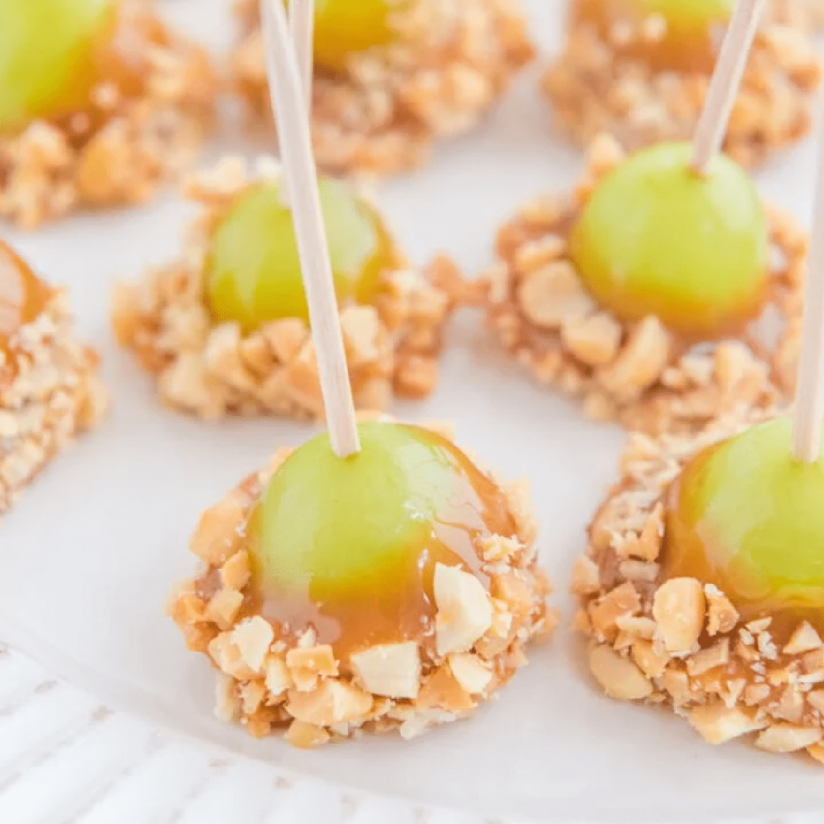 Mini caramel apple grape bites made with green grapes dipped in gooey caramel and chopped peanuts, secured with toothpicks and arranged on a white plate — fun and easy no-bake fall treat.