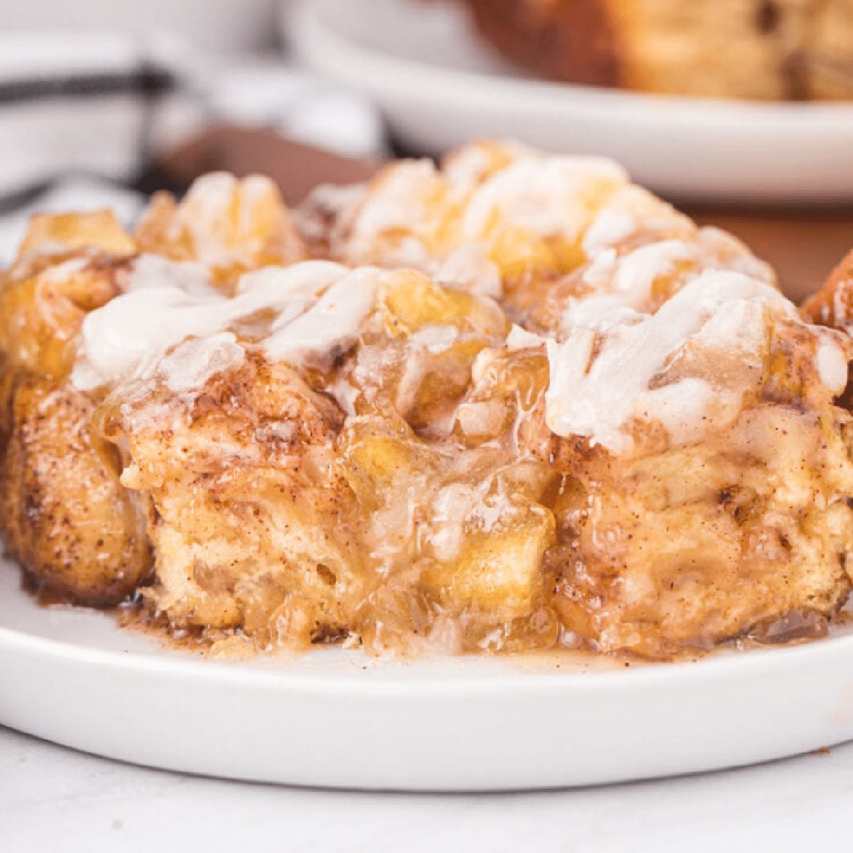 Close-up of gooey caramel apple pie cinnamon roll bake topped with creamy icing, served on a white plate.