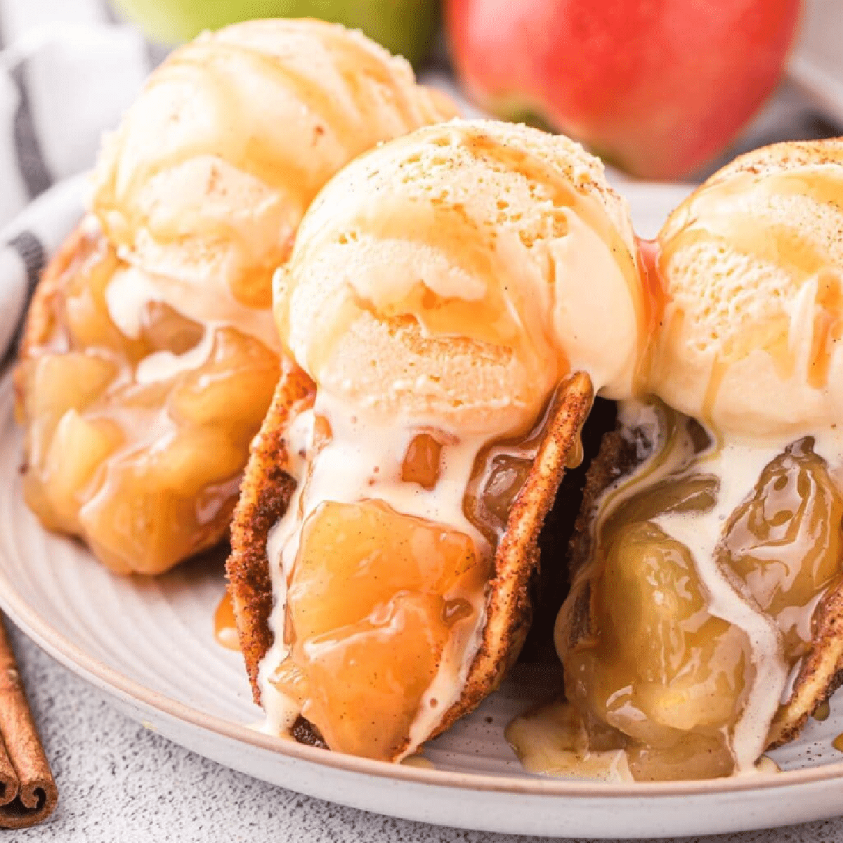 Three caramel apple pie tacos filled with gooey apple pie filling and topped with scoops of vanilla ice cream and drizzled caramel sauce, served on a plate with apples and cinnamon sticks in the background.