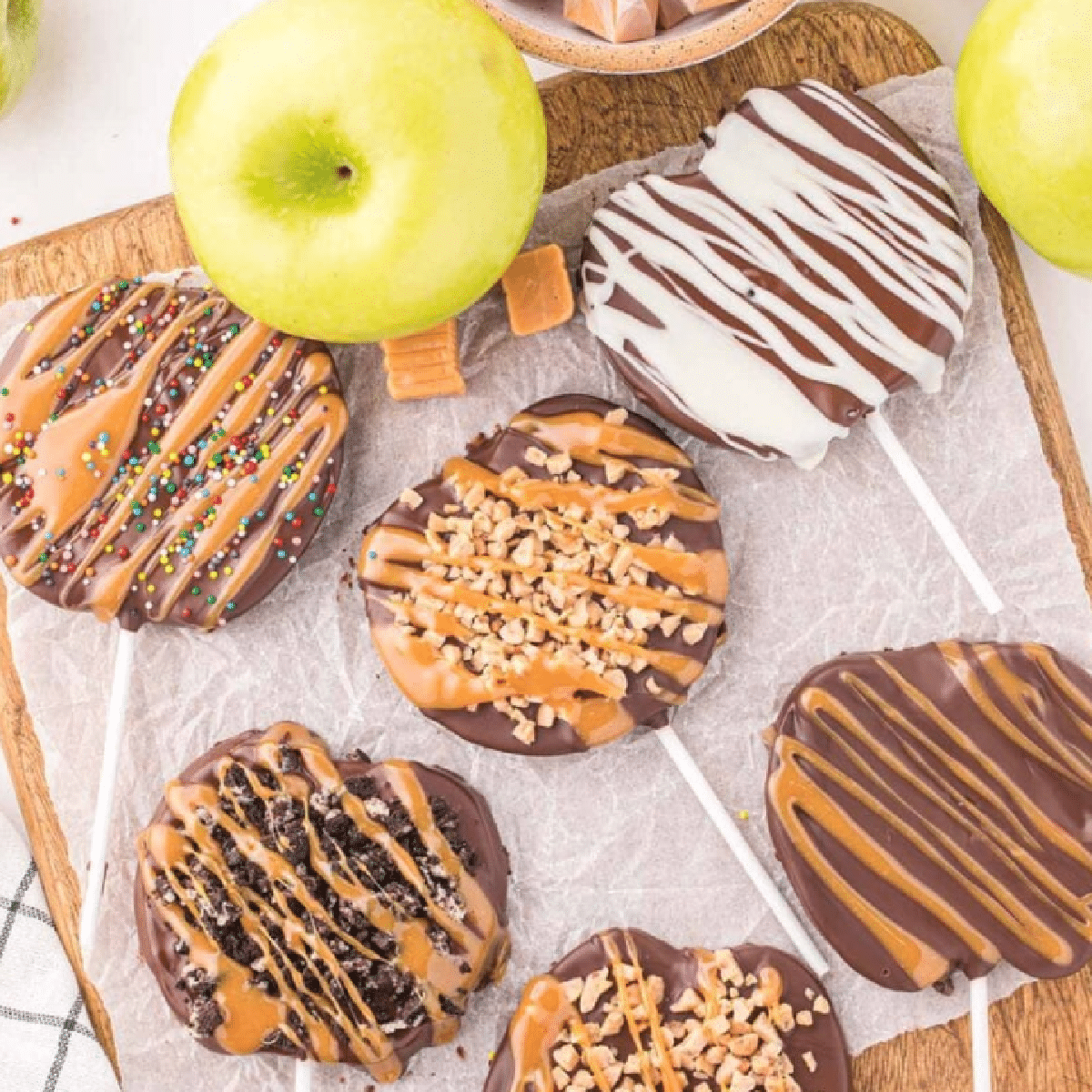 Chocolate-dipped caramel apple slices on sticks, topped with festive decorations like sprinkles, toffee bits, crushed cookies, and drizzle of caramel and white chocolate — easy no-bake treat served on parchment paper with fresh apples.