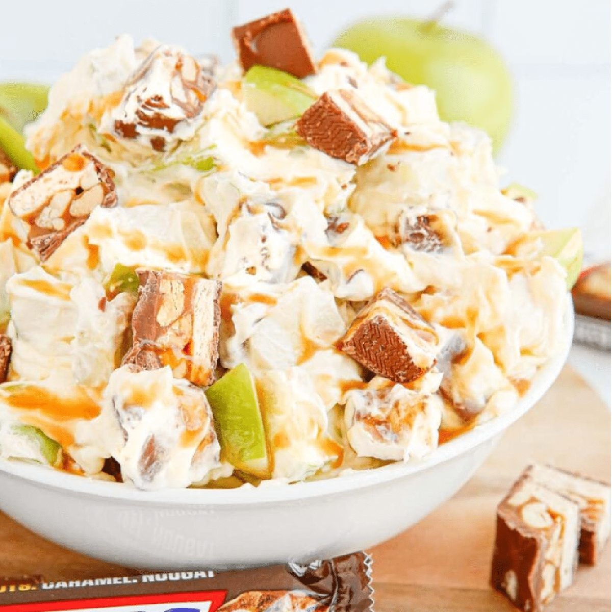 Close-up of a bowl filled with creamy caramel apple Snickers salad made with chopped green apples, whipped topping, and chunks of Snickers bars, drizzled with caramel sauce.
