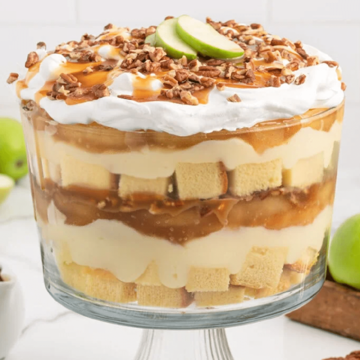 Glass trifle bowl filled with layers of cubed pound cake, creamy vanilla pudding, caramel apple pie filling, and whipped topping, finished with chopped pecans, caramel drizzle, and fresh green apple slices on top — elegant no-bake fall dessert.