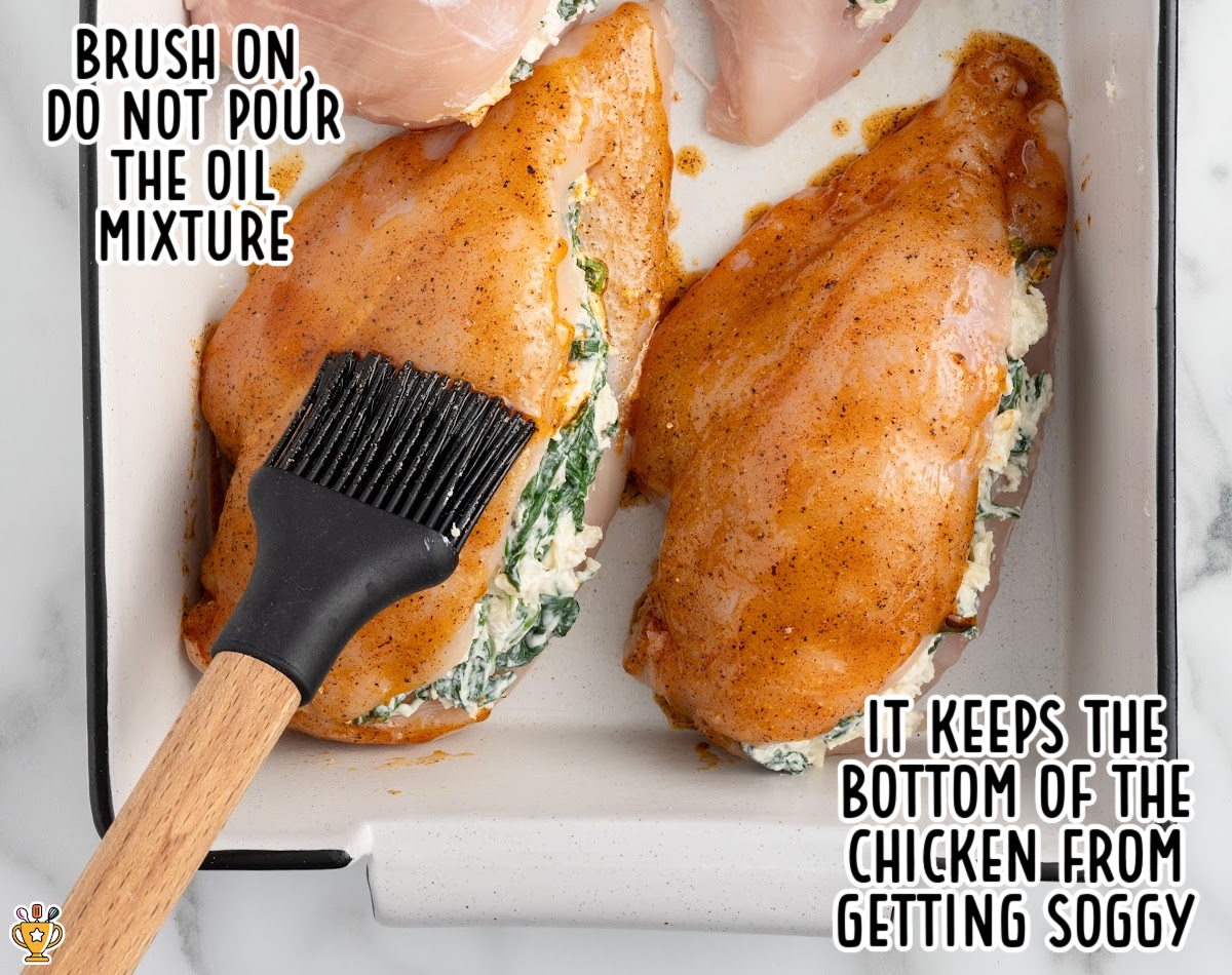 Unbaked spinach stuffed chicken breasts in a baking dish being brushed with a seasoned oil mixture to prevent the bottoms from getting soggy before baking.