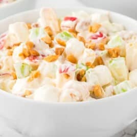 A white bowl filled with taffy apple salad, featuring a creamy mixture of chopped red and green apples, mini marshmallows, crushed peanuts, and pineapple tidbits, all coated in a whipped topping and pudding mixture. A few peanuts are scattered around the bowl on the white marble surface, with a spoon placed beside it.