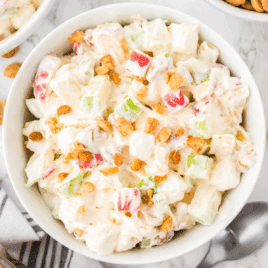 A close-up overhead view of a bowl filled with creamy taffy apple salad, featuring chunks of red and green apples, mini marshmallows, crushed peanuts, and a fluffy whipped topping dressing, served in a white bowl with a spoon nearby on a white marble surface.