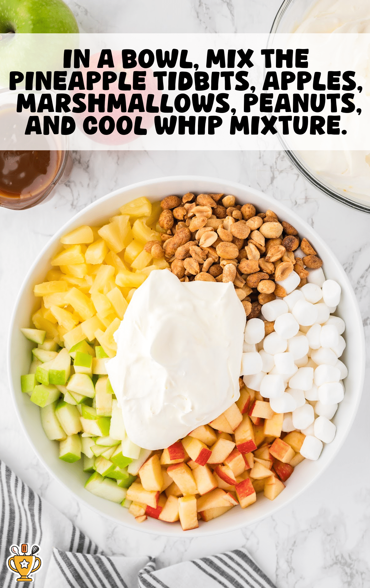 Overhead view of a large white mixing bowl filled with chopped green and red apples, crushed peanuts, mini marshmallows, pineapple tidbits, and a large dollop of Cool Whip in the center. A caramel sauce and Cool Whip bowl are partially visible above, with a green apple, red apple, and white background.