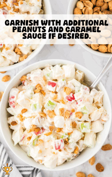A bowl of creamy taffy apple salad garnished with chopped peanuts and surrounded by additional bowls of salad and peanuts on a marble surface. The salad contains chunks of red and green apples, mini marshmallows, and a creamy mixture.